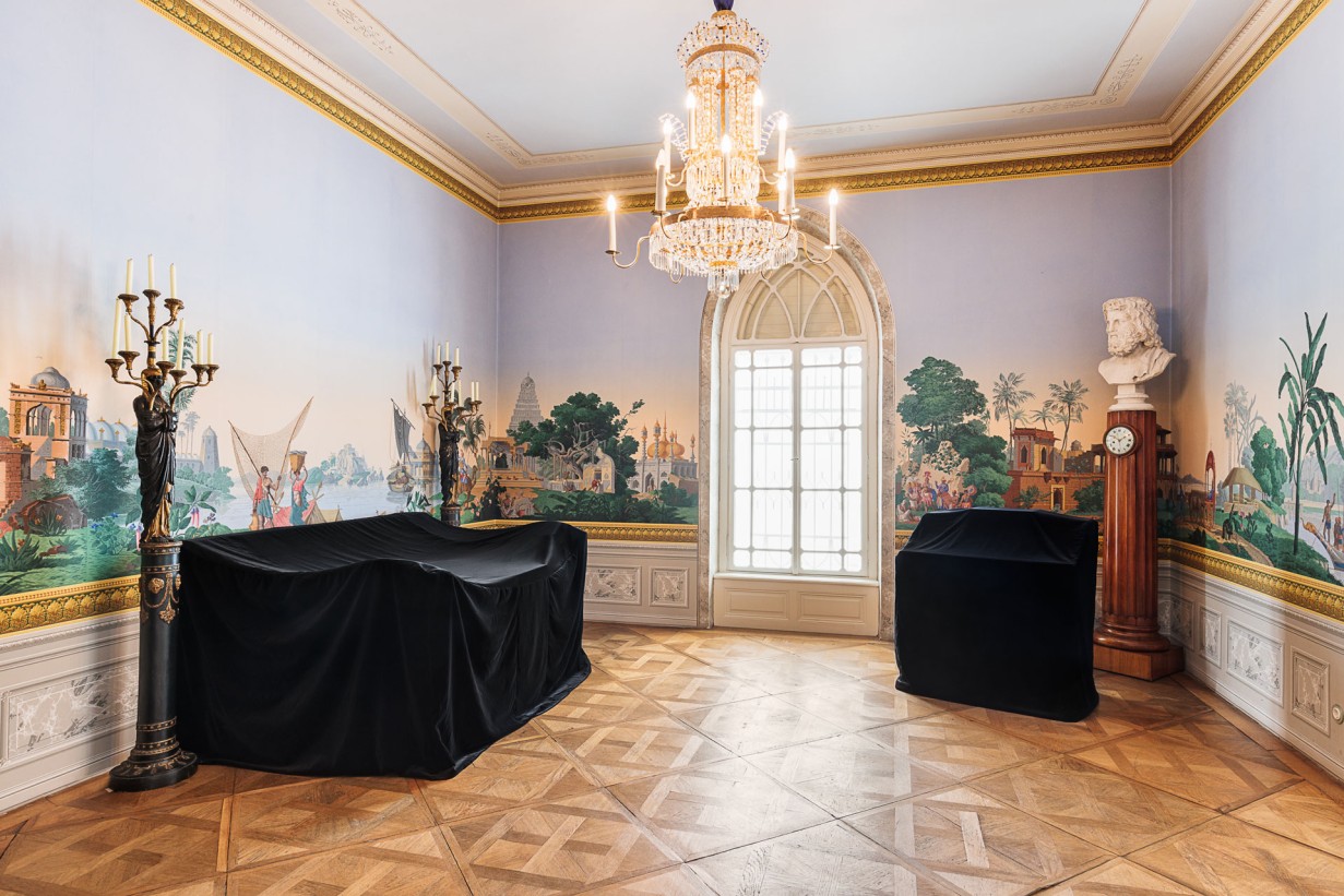 Frontal view of a room, on the walls a colorful wallpaper, a chandelier hangs from the ceiling, two pieces of furniture are covered with black fabric.