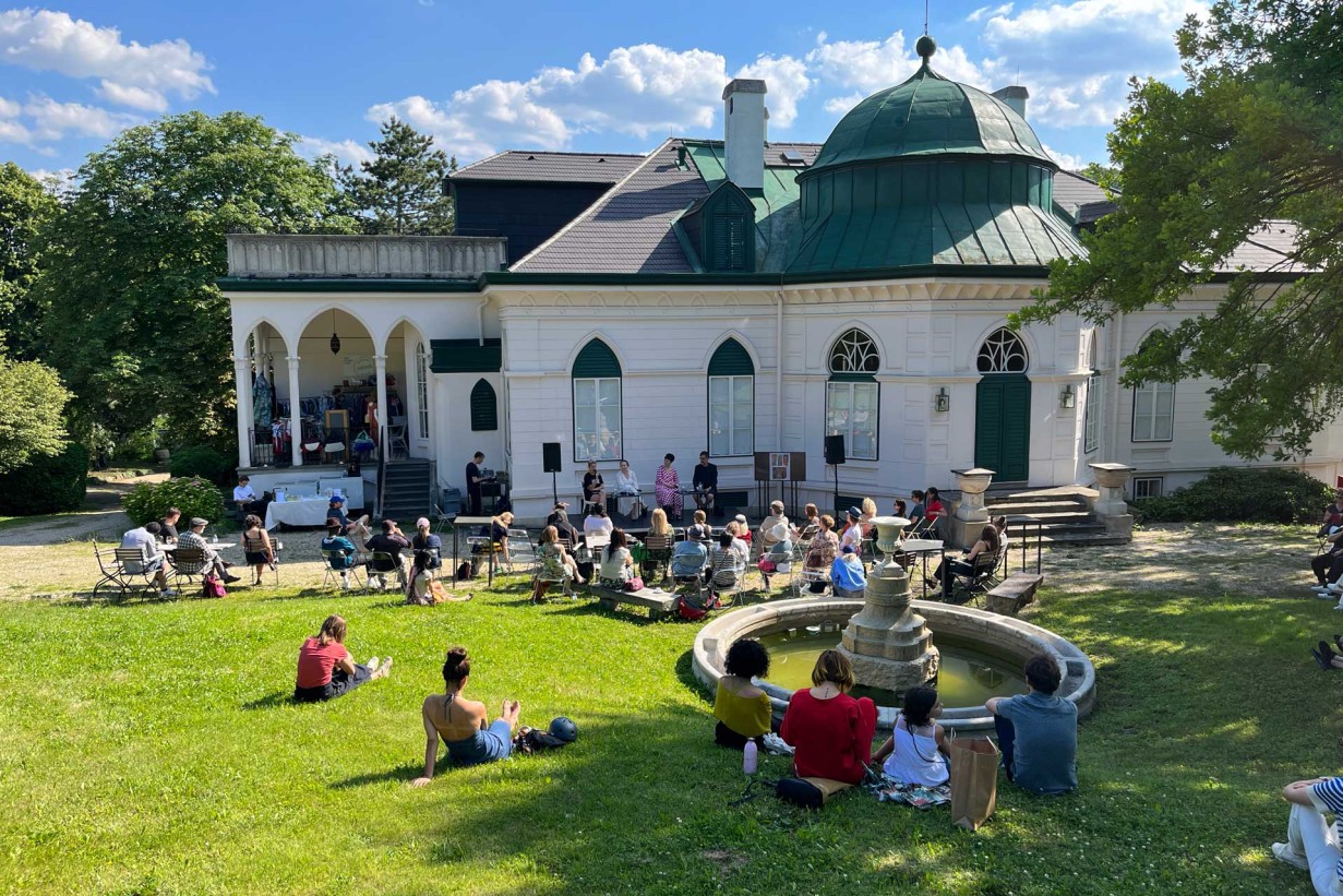 Schlosspark mit Besucher*innen, die im Gras sitzen. Schlössel im Hintergrund. 