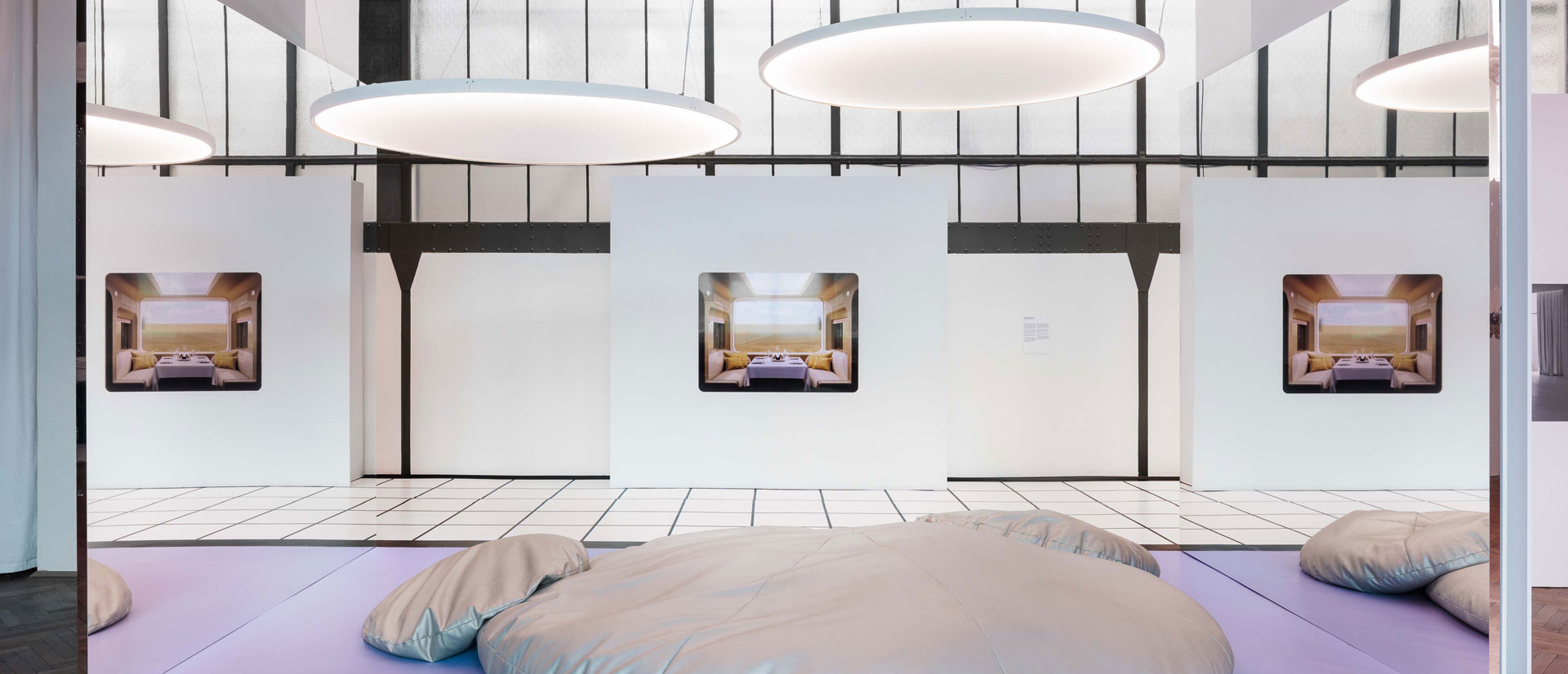 Ausstellungshalle, lila Boden, silber Sitzpolster, von der Decke hängen runde, flache Lampen