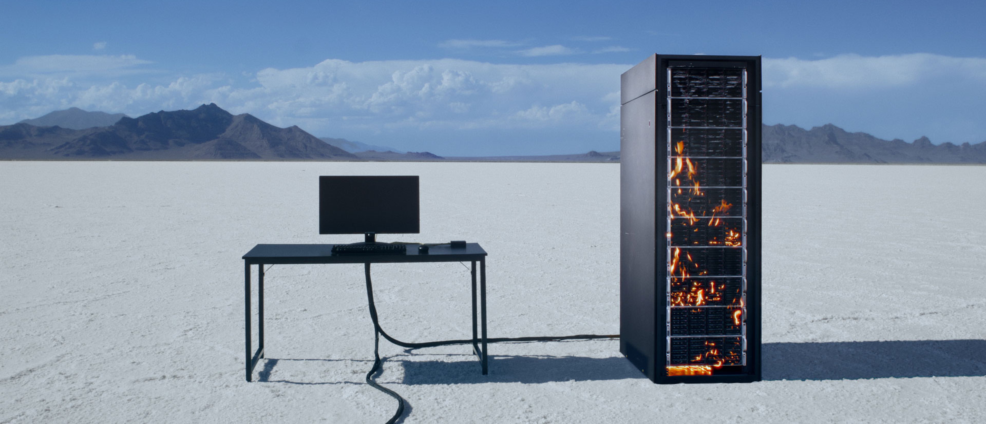 A computer screen on a desk, next to it a server tower in flames in a desert landscape.