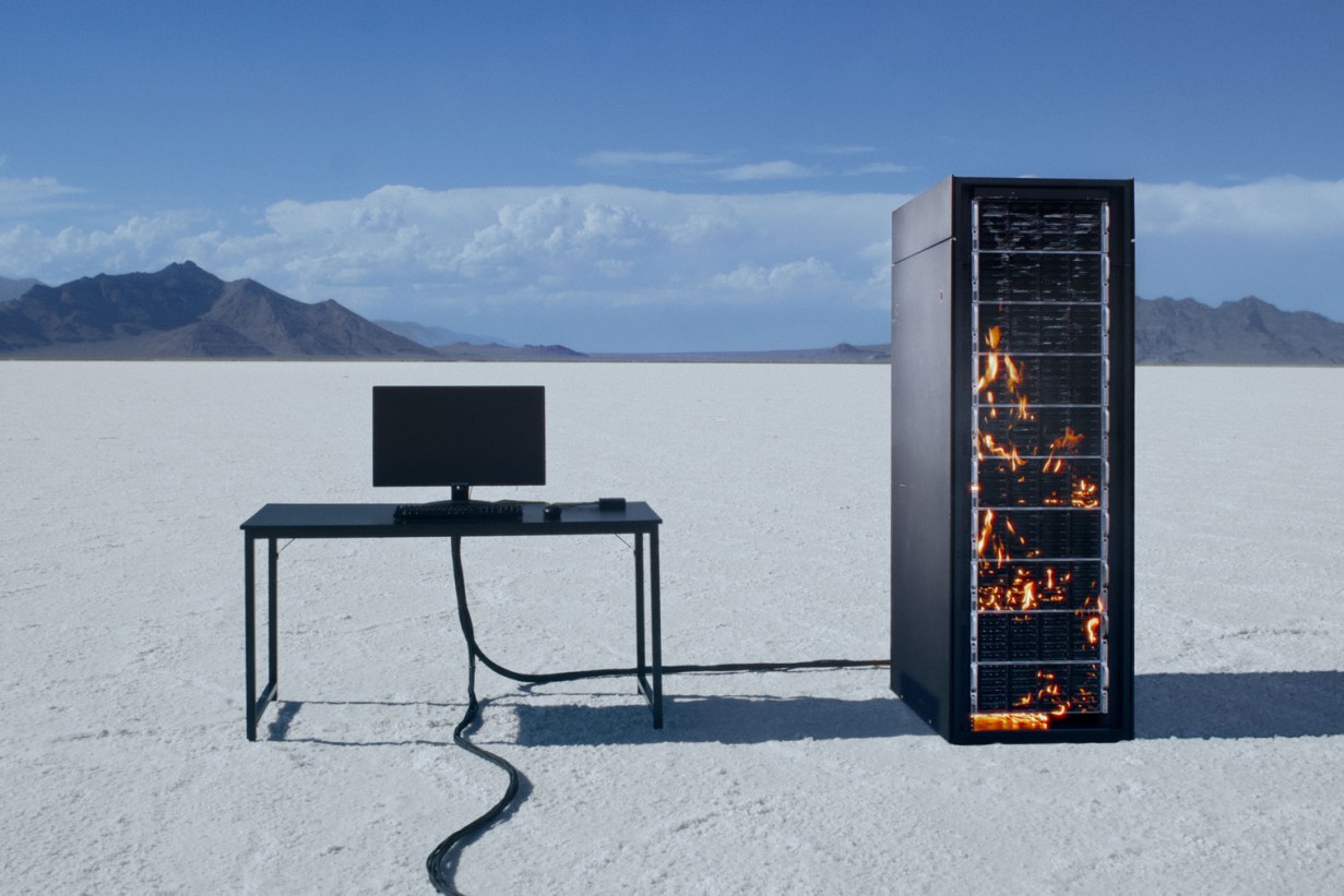 A computer screen on a desk, next to it a server tower in flames in a desert landscape.