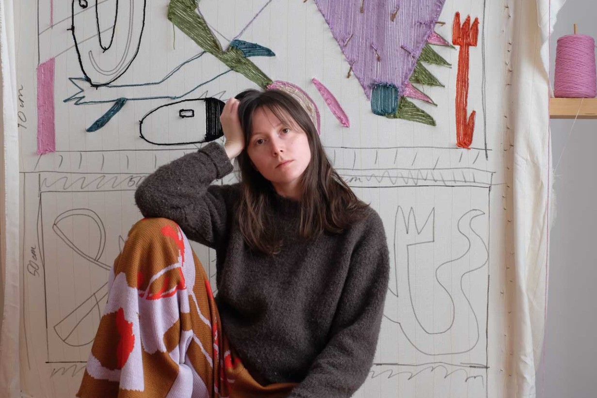 Junge weiße Frau sitzend vor einem getuffteten Teppich an der Wand.