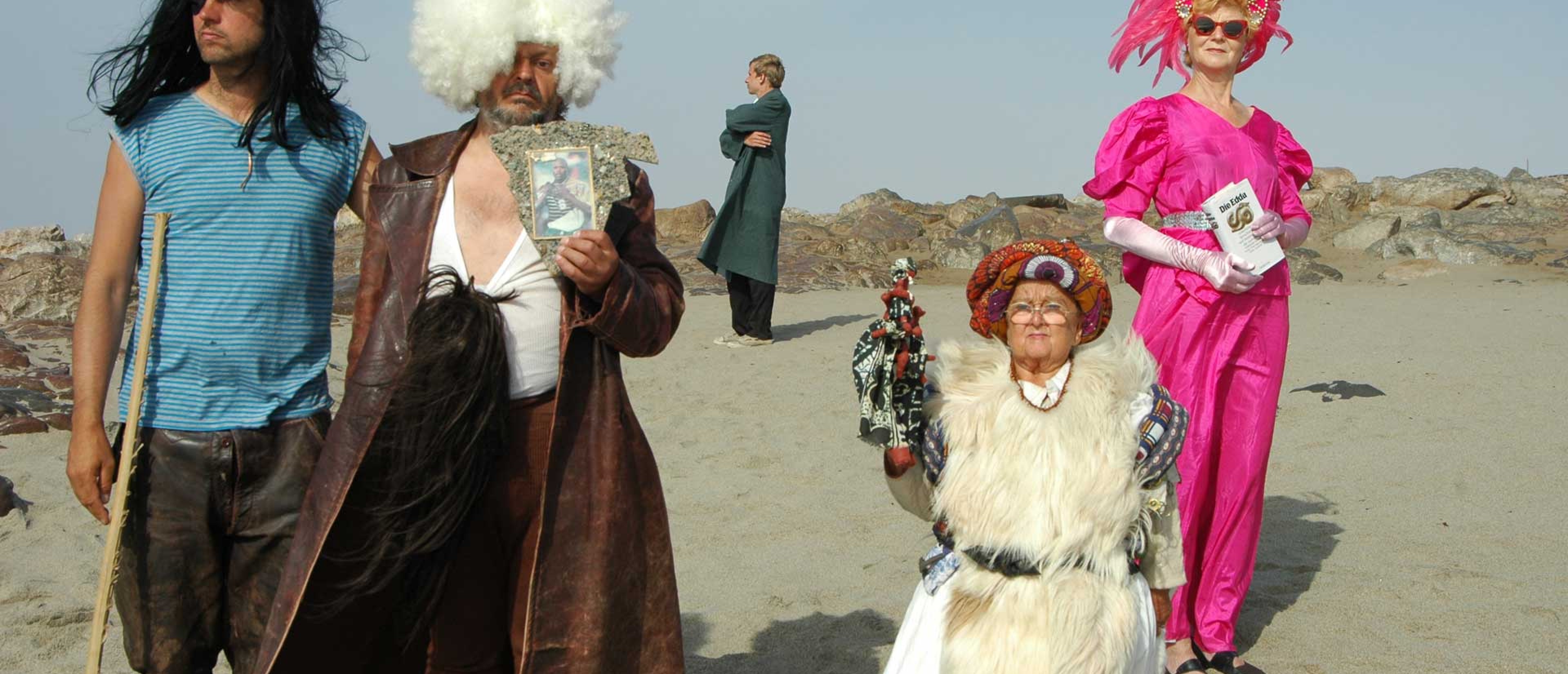 Five people wearing wigs stand in a desert-like setting.